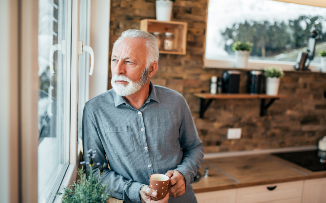 Nostalgic and melancholy senior man standing in the kitchen and looking through window, holding a cup of coffee or tea on a cold winter day.